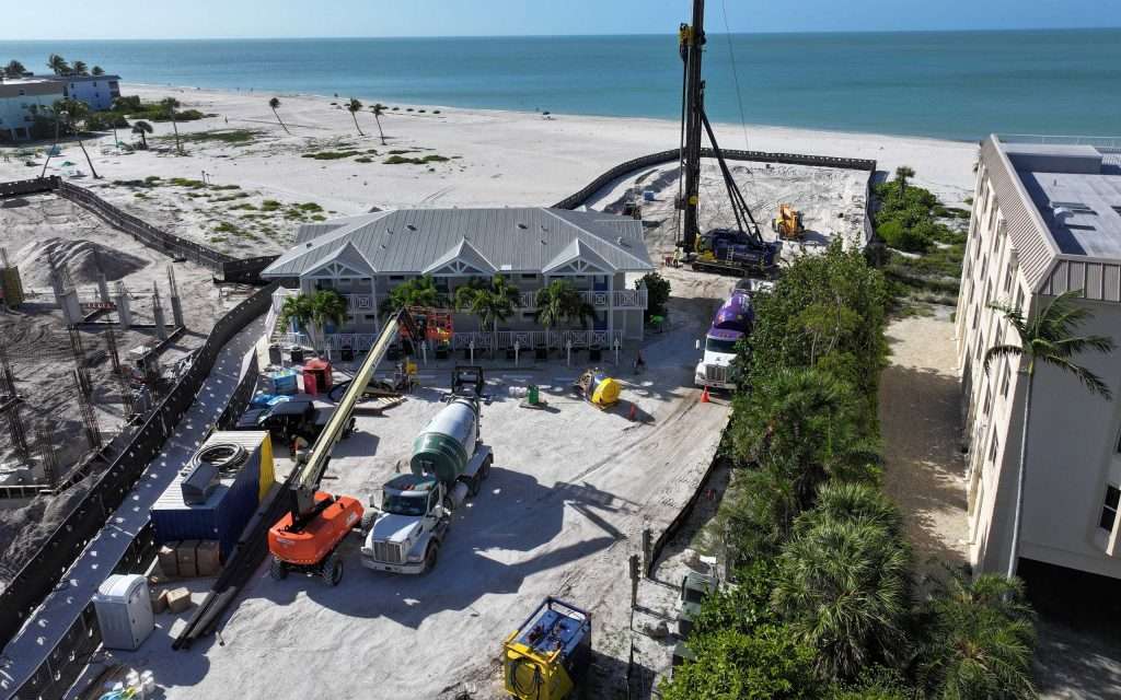 construction vehicles and equipment are positioned around a beachside building, with the ocean and sandy shoreline visible in the background.