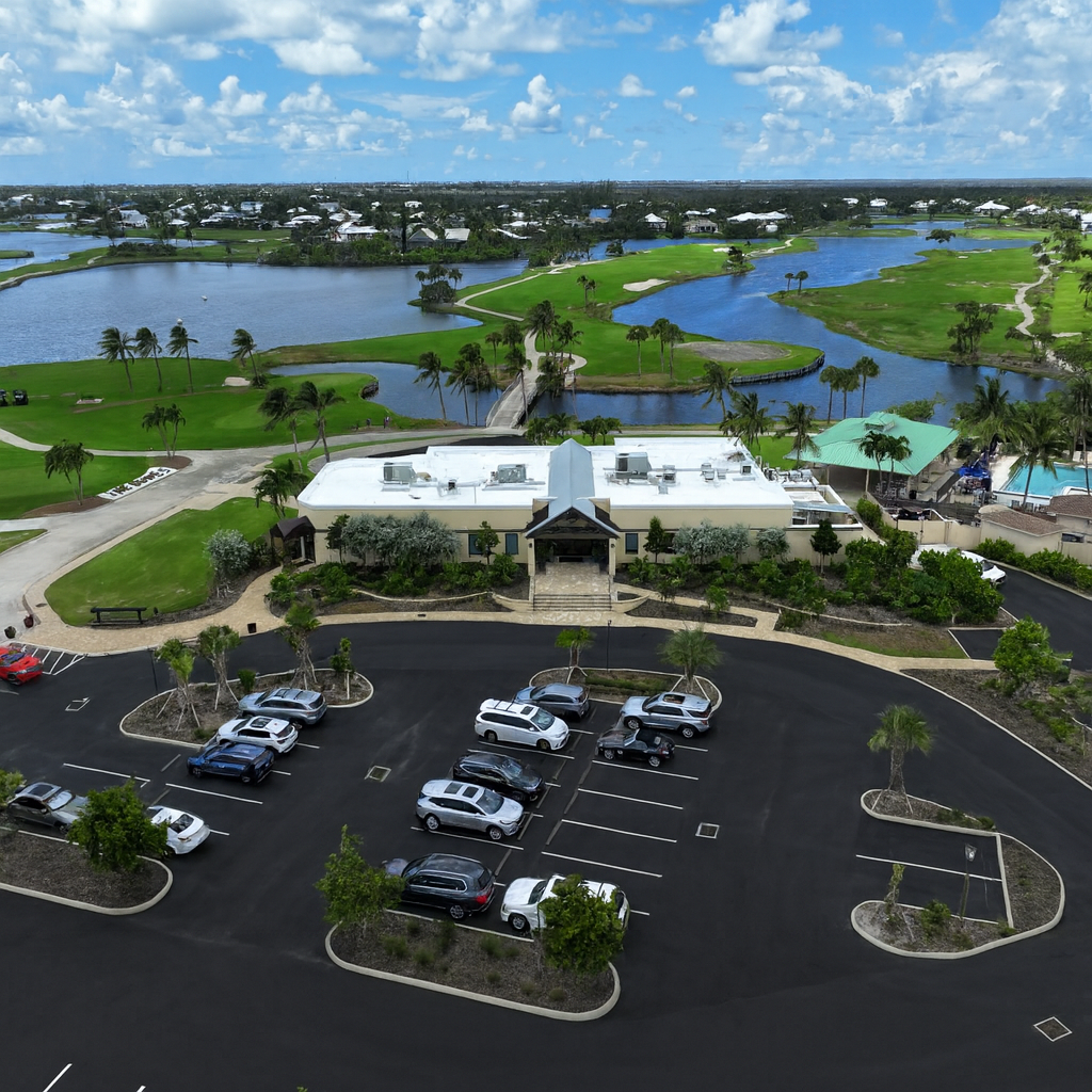 Aerial view of a parking lot with cars, a modern building, golf course, water features, and residential homes under a partly cloudy sky.