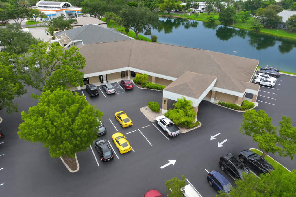 Aerial view of a commercial building with a parking lot, several parked cars, trees, and a pond in the background.