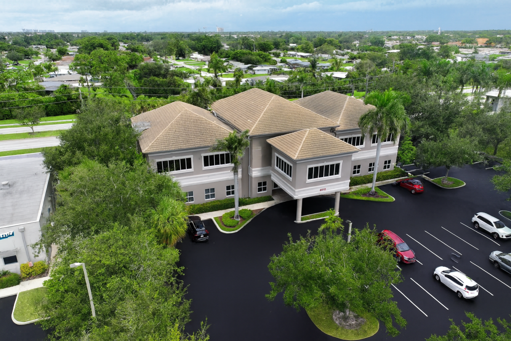Aerial view of a two-story office building with a tan roof, surrounded by trees and parked cars, located in a suburban area.