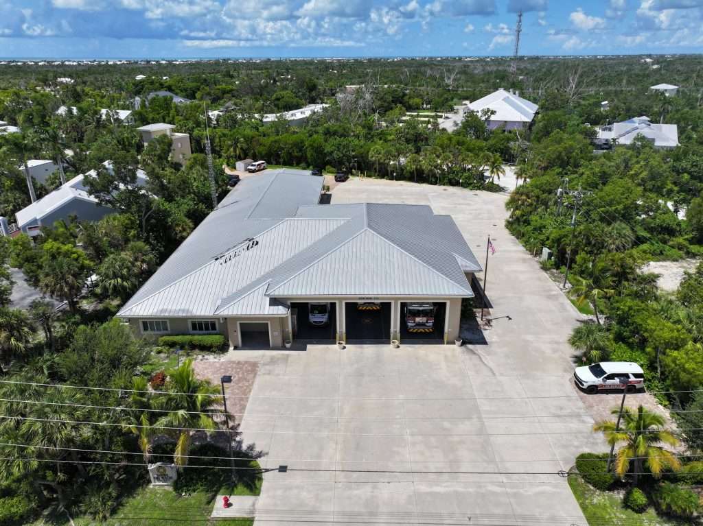 Aerial view of a fire station with three vehicle bays, a flagpole, parked vehicles, and surrounding trees and buildings.