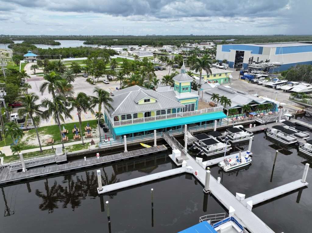 Aerial view of a waterfront marina with a turquoise building, palm trees, parked boats, and docks under a cloudy sky.