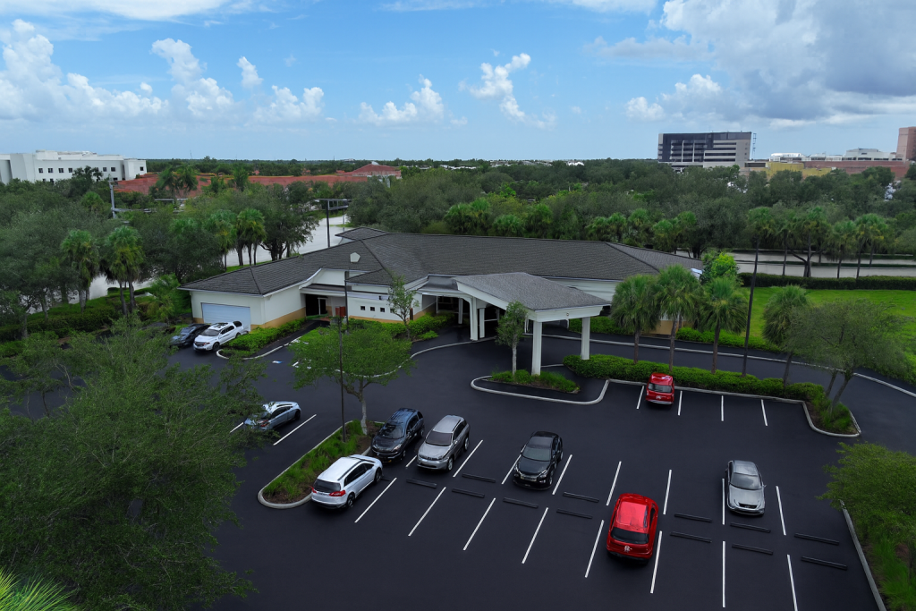 A single-story building with a covered entrance is surrounded by palm trees and a parking lot with several cars, under a partly cloudy sky.