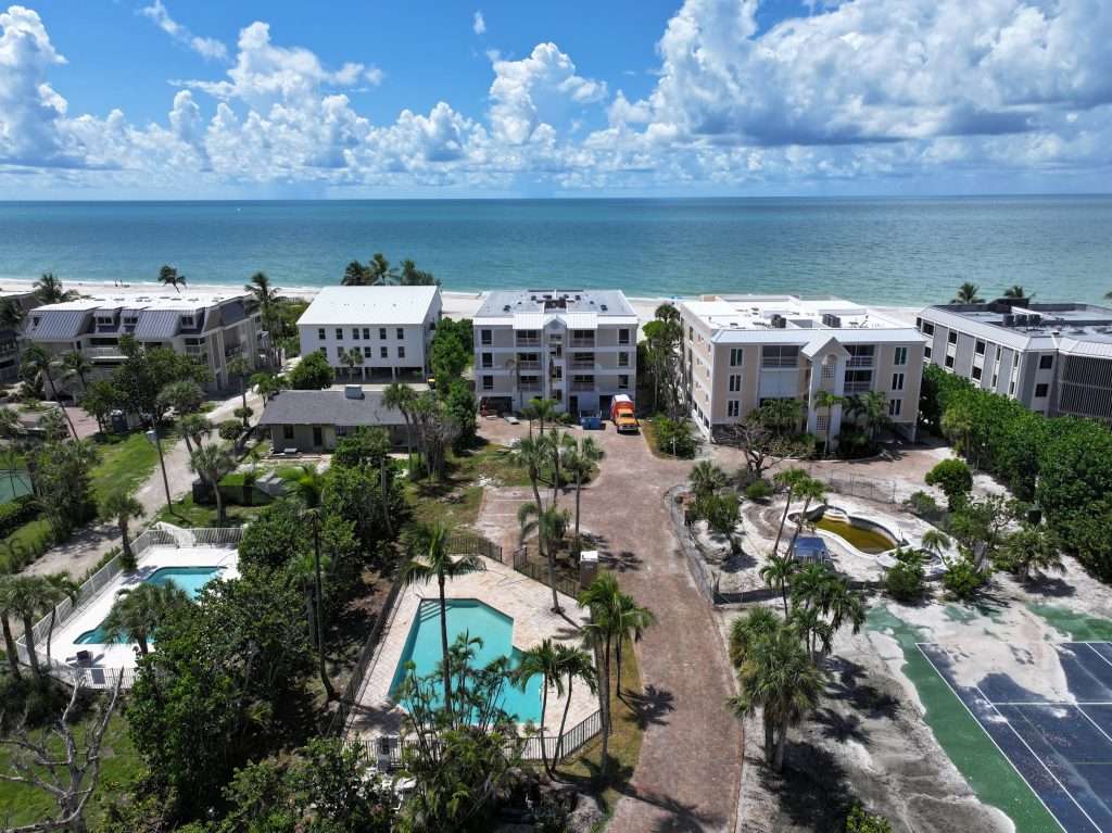 Aerial view of a coastal residential complex with multiple buildings, swimming pools, palm trees, and tennis courts near the ocean under a partly cloudy sky.