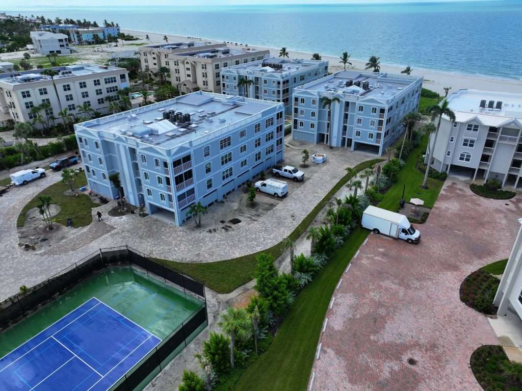 Aerial view of apartment complexes near the beach with trucks parked outside and a tennis court in the foreground.