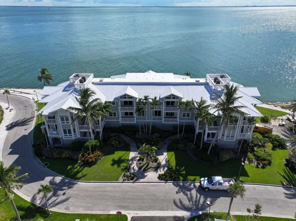 Aerial view of a multi-story coastal condominium building with a white roof, surrounded by palm trees and landscaping, next to a body of water.