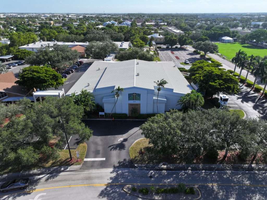 Aerial view of a large, white, single-story building surrounded by trees, with parking areas and a road in the foreground. Residential neighborhood and green spaces in the background.