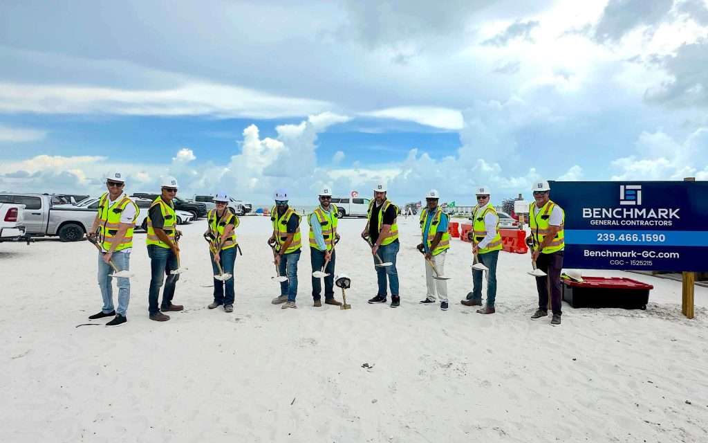 nine people in safety gear and hard hats stand in a row holding shovels on a sandy construction site, with vehicles and a benchmark contractors sign in the background.