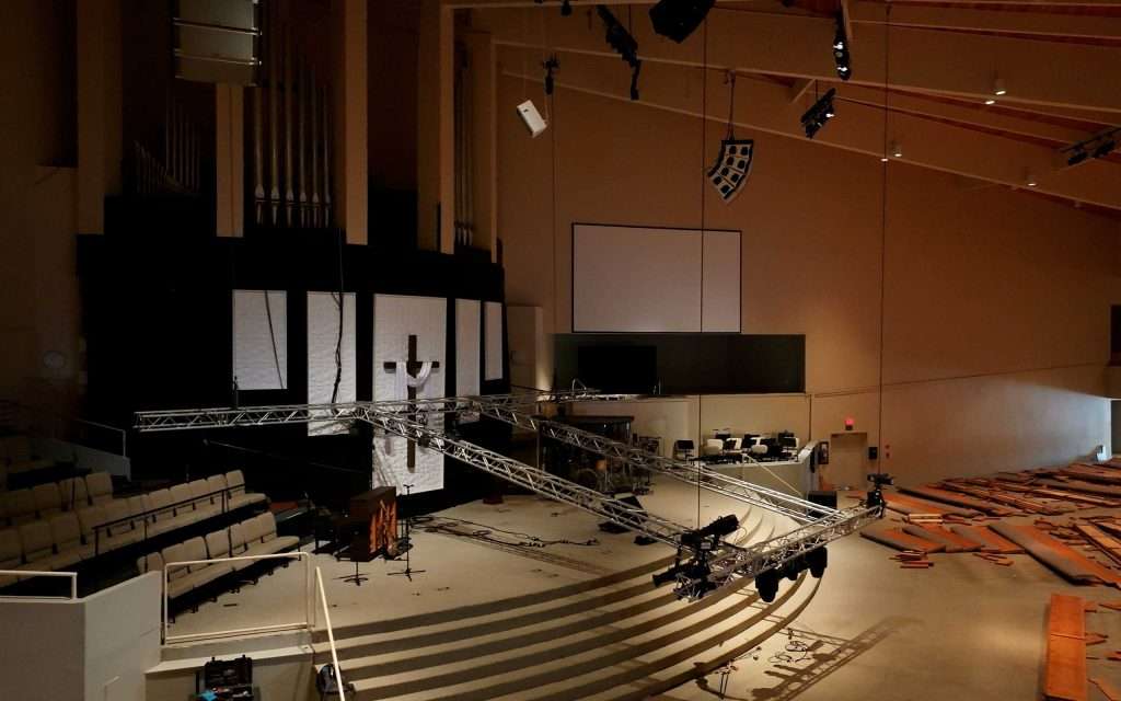 empty church auditorium with lighting equipment set up over the stage, organ pipes on the left, and wooden pews stacked on the right side of the room.
